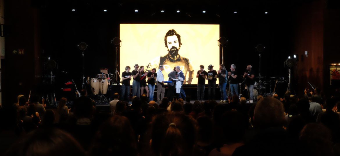 Imagen concierto jarabe de palo homenaje a Pau Donés siluetas de artistas en el escenario con una imagen de Pau Donés en pantalla